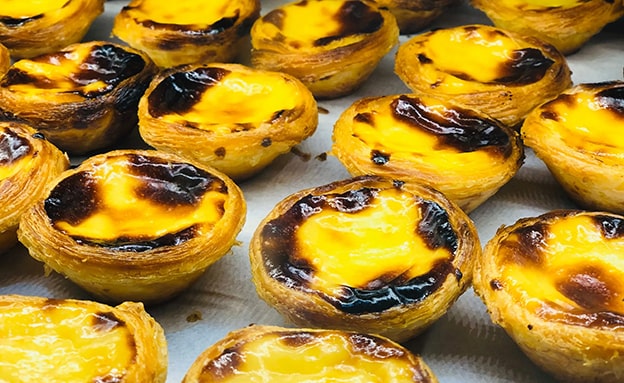 Pastel de Nata (Portekiz)

18. yüzyılda Lizbon’daki Jerónimos Manastırı’nda doğan pastel de nata, çıtır hamur içinde krema dolgulu bir turtadır. Rahipler, yumurta sarılarını değerlendirmek için bu tatlıyı geliştirmiş. Yüksek ısıda pişirilen tatlı, hafif yanık yüzeyiyle ünlüdür ve tarçınla servis edilir. Zamanla Portekiz’in ulusal sembolü haline gelmiştir.